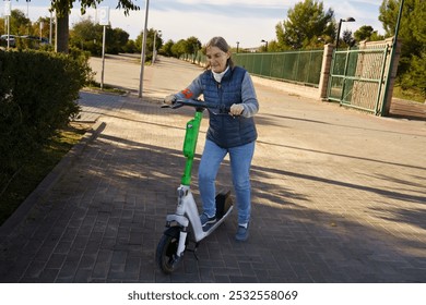 An elderly woman riding an electric scooter on a paved path in a park-like setting. She is wearing a blue vest and jeans, smiling as she enjoys her ride. The background features greenery and a gated a - Powered by Shutterstock