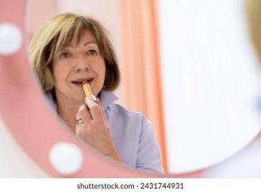 An elderly woman with short brown hair applying lipstick in a mirror. She is wearing a blue blouse and has a serene expression. The background is soft and out-of-focus, creating a peaceful atmosphere. - Powered by Shutterstock