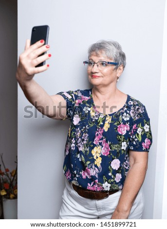 Elderly woman with short gray hair taking selfies and pictures with her smart phone in front of light gray wall. The mature woman is holding the phone in the right hand looking at screen