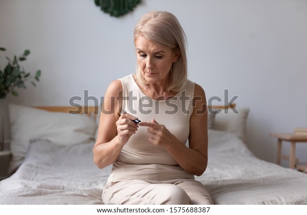 Elderly woman sit on bed in morning holding\
glucose meter lancets needles pricks skin obtain blood sample,\
diabetic patient easily do check up control of blood sugar levels\
at home, diabetes\
concept