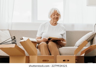 elderly woman sits on a sofa at home with boxes. collecting things with memories albums with photos and photo frames moving to a new place cleaning things and a happy smile. Lifestyle retirement. - Powered by Shutterstock