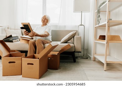 elderly woman sits on a sofa at home with boxes. collecting things with memories albums with photos and photo frames moving to a new place cleaning things and a happy smile. Lifestyle retirement. - Powered by Shutterstock