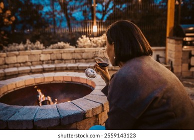 Elderly woman sitting alone by fire pit, drinking glass of red wine. - Powered by Shutterstock