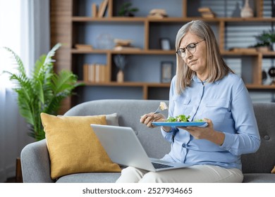 Elderly woman sitting on couch, eating salad, looking dissatisfied, working on laptop at home. Reflects feelings of frustration with healthy diet, multitasking, home office. No background features. - Powered by Shutterstock