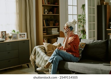 Elderly woman sitting on couch while holding smartphone, engaged in conversation. Cozy living room with bookshelves, photos and indoor plants creating warm atmosphere - Powered by Shutterstock