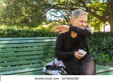 Elderly woman sitting on park bench outdoors coughing into elbow and holding tissue - coronavirus concept (selective focus)