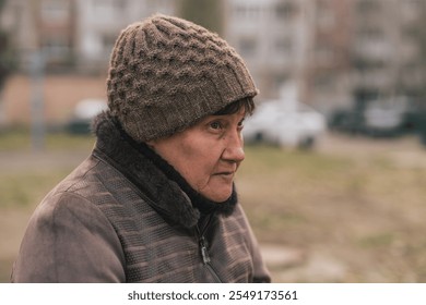 Elderly woman sitting on a park bench wearing a knitted hat during a cool autumn day in an urban setting. - Powered by Shutterstock