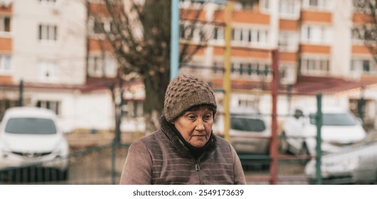 Elderly woman sitting on a park bench wearing a knitted hat during a cool autumn day in an urban setting. - Powered by Shutterstock