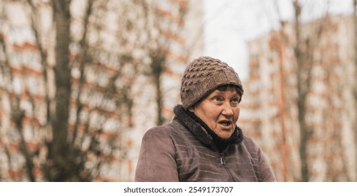 Elderly woman sitting on a park bench wearing a knitted hat during a cool autumn day in an urban setting. - Powered by Shutterstock