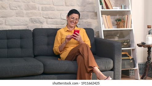 Elderly woman sitting on the sofa at home, reading the news on her cell phone. - Powered by Shutterstock