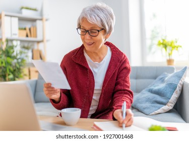 Elderly Woman, Sitting On The Sofa With A Paper Receipt In Her Hands, Are Calculating Expenses, Managing The Family Budget.