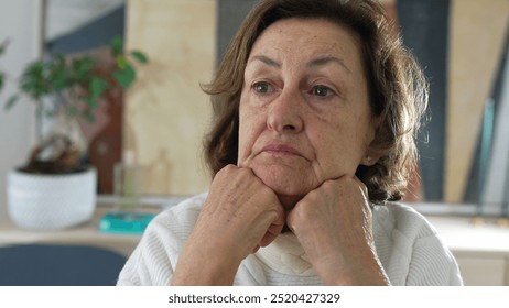 Elderly woman sitting at a table, resting her chin on both hands, looking down in contemplation. a quiet, reflective moment in a serene home environment, emphasizing solitude - Powered by Shutterstock