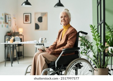 Elderly woman sitting in wheelchair in modern office holding tablet and looking at camera, with plants and office supplies in background, creating a professional atmosphere - Powered by Shutterstock