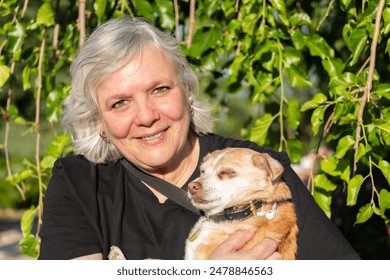 Elderly woman smiling and holding her small dog in a sunlit garden. She smiles warmly as she holds her small light brown dog in a sunlit garden. - Powered by Shutterstock