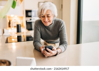 Elderly Woman Speaking To A Smart Speaker In Her Kitchen. Happy Senior Woman Using A Virtual Assistant To Perform Household Tasks In Her Smart Home.