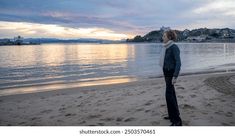 Elderly woman standing alone on a sandy beach at sunset, looking at the horizon - Powered by Shutterstock