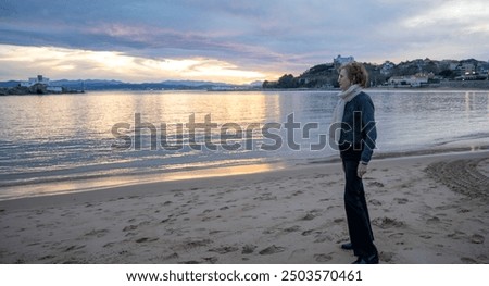 Elderly woman standing alone on a sandy beach at sunset, looking at the horizon