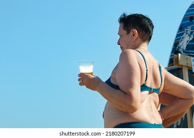 An Elderly Woman Stands With A Glass Of Beer On The Beach. Sunny Day, Summer.