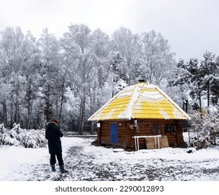 An elderly woman stands at the round house of the ail under the trees in the snow in winter in the Altai in Siberia.