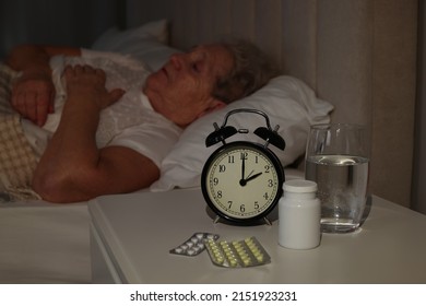 Elderly Woman Suffering From Insomnia In Bed At Home, Focus On Pills And Alarm Clock
