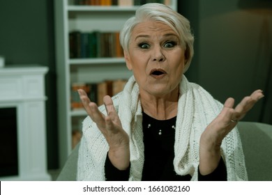 Elderly Woman Surprised. She Raised Her Eyebrows In Surprise And Raised Her Hands While Sitting In A Chair In Cozy Interior At Home. Toned Image