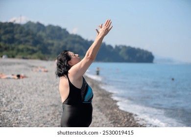 An elderly woman in a swimsuit does sports and breathing exercises on the beach. - Powered by Shutterstock