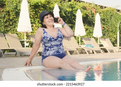 Elderly Woman In Swimsuit Sits On The Side Of The Pool And Put Her Feet In The Water And Talking On A Mobile Phone. Mature Woman In Panama Is Relaxing In Hotel. Woman Smiles And Looks At The Camera.