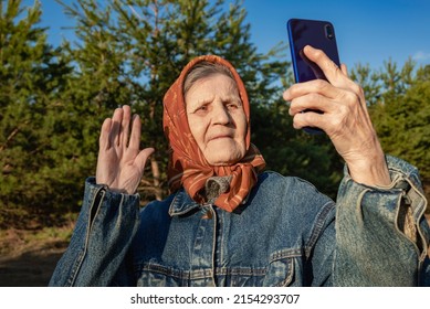 An Elderly Woman Takes Photo Of Selfie On Mobile Phone While Walking In A Pine Forest. Grandma Communicates Via Video Communication In A Smartphone. Modern Old Lady In A Denim Jacket And A Headscarf.