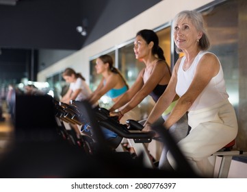 Elderly woman taking indoor cycling class at fitness center, doing cardio riding bike - Powered by Shutterstock