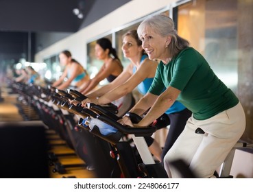 Elderly woman taking indoor cycling class at fitness center, doing cardio riding bike - Powered by Shutterstock