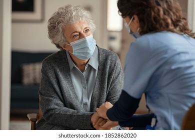 Elderly woman talking with a doctor while holding hands at home and wearing face protective mask. Worried senior woman talking to her general practitioner visiting her at home during virus epidemic. 