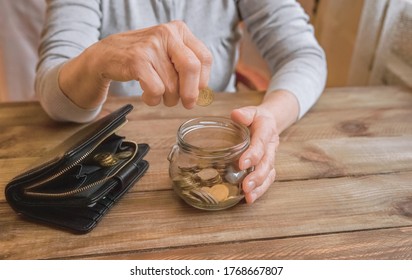 Elderly Woman Throws A Coin Into A Jar, Counting. Old Wrinkled Hand Holding Jar With Coins, Empty Wallet, Wooden Background. Saving Money For Future, Retirement Fund, Pension, Poorness, Need Concept.