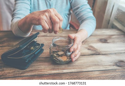 Elderly Woman Throws A Coin Into A Jar, Counting. Saving Money For Future, Retirement Fund, Pension, Poorness, Need Concept. Old Wrinkled Hand Holding Jar With Coins, Empty Wallet, Wooden Background. 