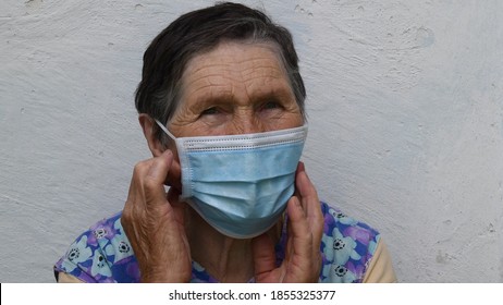 Elderly Woman Touches Mask By Hands. Old Lady Wears Face Mask To Protect From Viruses And To Prevent Infestation. Mask At Wrinkled Female Face Of 80 Year Old Woman