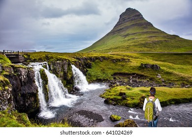 Elderly Woman -  Traveler With A Large Backpack Admiring The Powerful Jets Of The Waterfall Kirkjoufellfoss. Concept Of Exotic And Extreme Tourism. Summer In Iceland