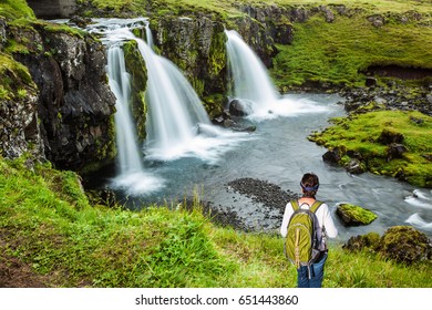 Elderly Woman -  Traveler With A Large Backpack Admiring The Powerful Jets Of The Waterfall Kirkjoufellfoss. Summer In Iceland. Concept Of Exotic And Extreme Tourism 