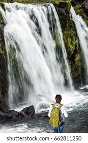 Elderly Woman -  Traveler With A Large Backpack Admiring The Powerful Jets Of The Waterfall Kirkjoufellfoss. Summer In Iceland. Concept Of Exotic And Extreme Tourism 