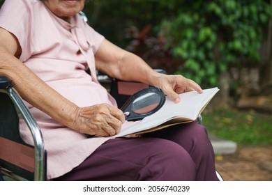 Elderly Woman Using A Magnifying Glass To Read On Wheelchair.