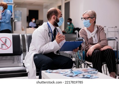 Elderly Woman Using Wheelchair Talking To Geriatrician At Medical Clinic Waiting Room. Various Ethnicities People Wearing Masks In Busy Sanatorium. Doctor Explaining Treatment To Caucasian Patient.