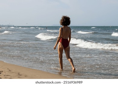 An elderly woman is walking along the shore of a sandy beach, wearing a red one-piece swimsuit. The serene beach setting and her relaxed demeanor capture a moment of leisure and enjoyment by the sea. - Powered by Shutterstock