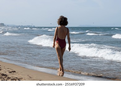 An elderly woman is walking along the shore of a sandy beach, wearing a red one-piece swimsuit. The serene beach setting and her relaxed demeanor capture a moment of leisure and enjoyment by the sea. - Powered by Shutterstock