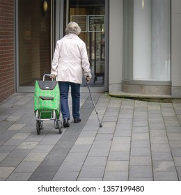 Elderly Woman With Walking Stick And Green Patterned Shopping Scooter Goes To A Double-winged Door