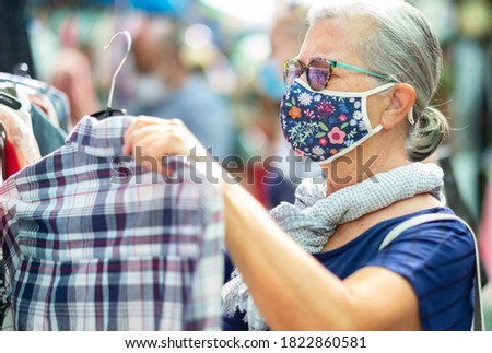 Elderly woman wearing a medical mask due to coronavirus while shopping at the flea market. Concept of buying cheap used clothes