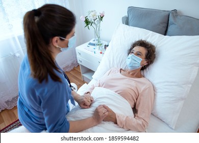 Elderly Woman Wearing Protection Face Mask Looking At Nurse Leaning Over. Top View Of Home Caregiver With Face Mask Holding Hands Of Female Senior Patient Lying In Bed At Nursing Home.