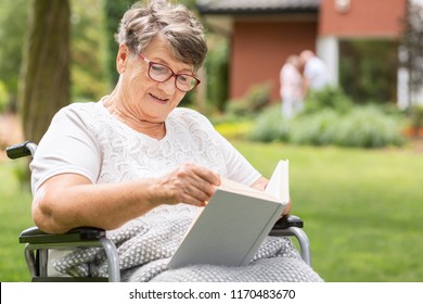 An Elderly Woman In A Wheelchair Reading A Book During Summer Day Outside In The Garden Of A Care Home For Seniors. Blurred Background.