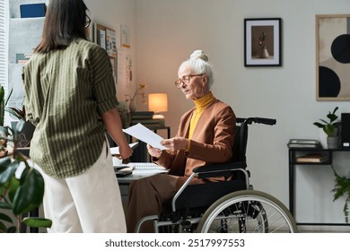 Elderly woman in wheelchair receiving document from colleague in office. Desk with laptop, lamp, and plant visible, and walls adorned with abstract art and pinned notes - Powered by Shutterstock