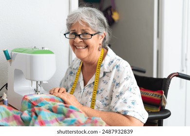 Elderly woman in a wheelchair smiling while sewing fabric on a sewing machine with a measuring tape around her neck. Concept of adaptive sewing. - Powered by Shutterstock