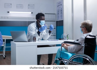 Elderly Woman In Wheelchair Wearing Virus Protection Facemask Having An Appointment With Clinic Specialist. Physician And Patient Talking About Pandemic Health Risks And Prevention Through Vaccination