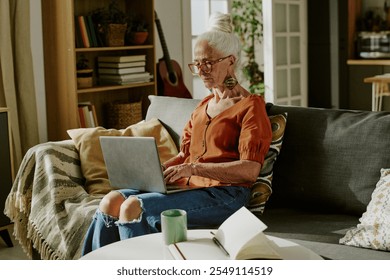 Elderly woman with white hair and glasses sitting on sofa using laptop in cozy living room having a green mug placed on table near her - Powered by Shutterstock