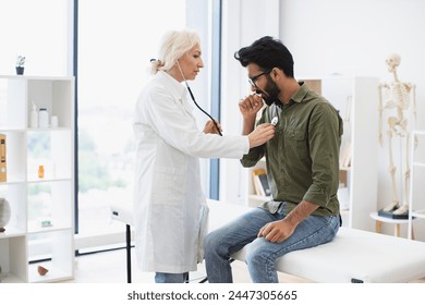 Elderly woman in white lab coat checking lungs and heart via stethoscope. Serious bearded man in everyday wear visiting family doctor for full examination in general practice. - Powered by Shutterstock